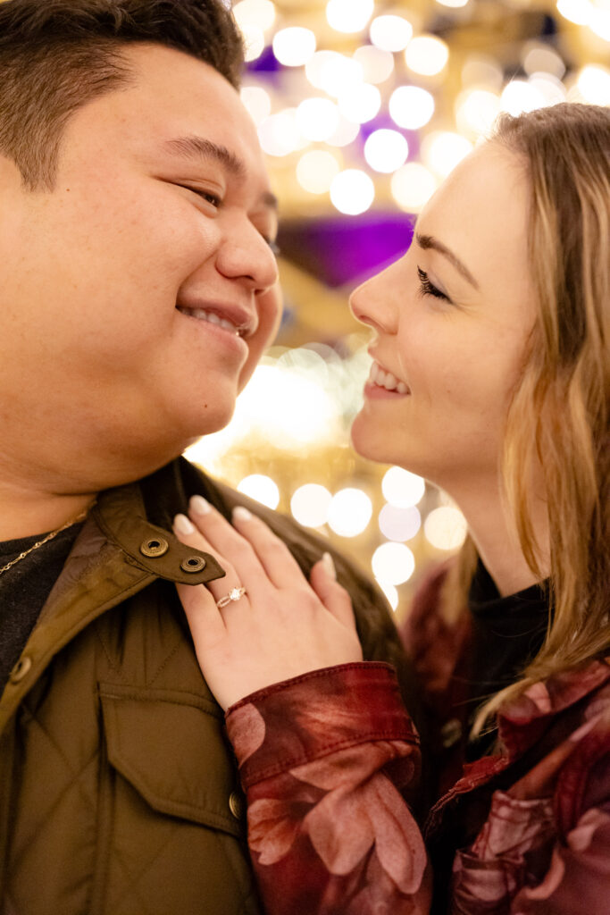 Engagement ring close-up on shoulder with couple looking into each other’s eyes, holiday lights in background