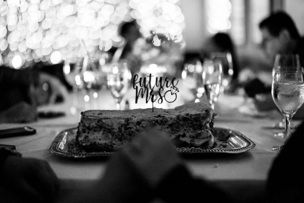Black and white photo of engaged couple with dessert that says "Future Mrs" surrounded by family