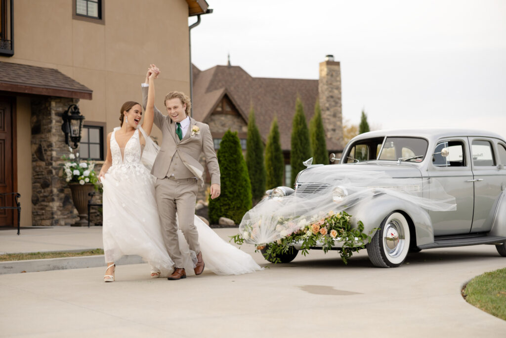 Bride and groom posing in front of a vintage car and elegant wedding venue, veil flying off in the wind