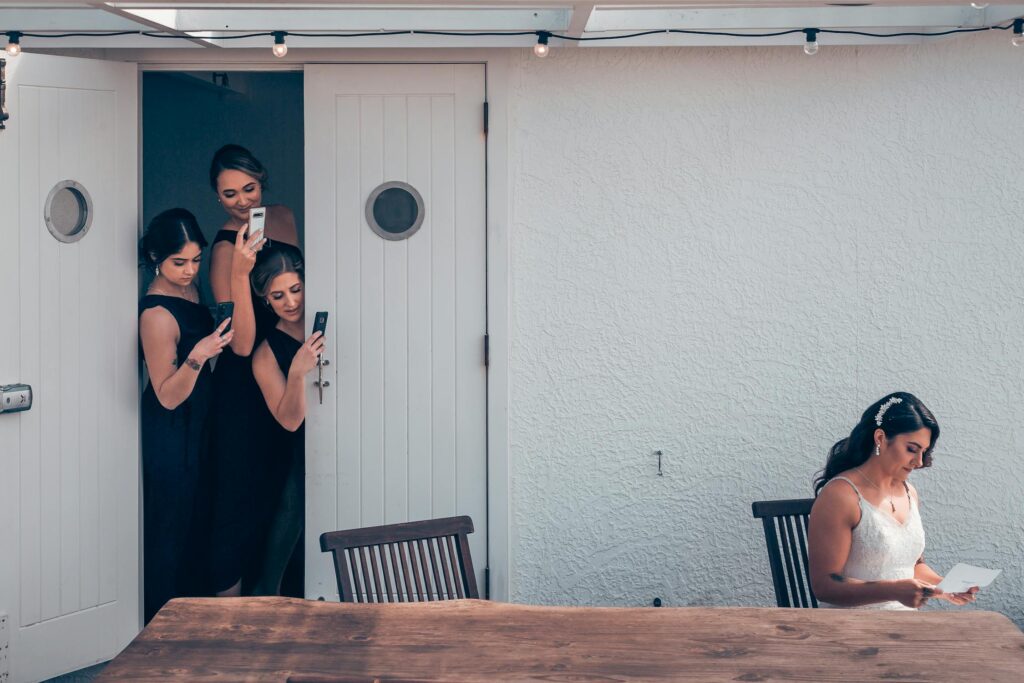 Bride reading a private letter from the groom while bridesmaids photograph through a cracked door from behind